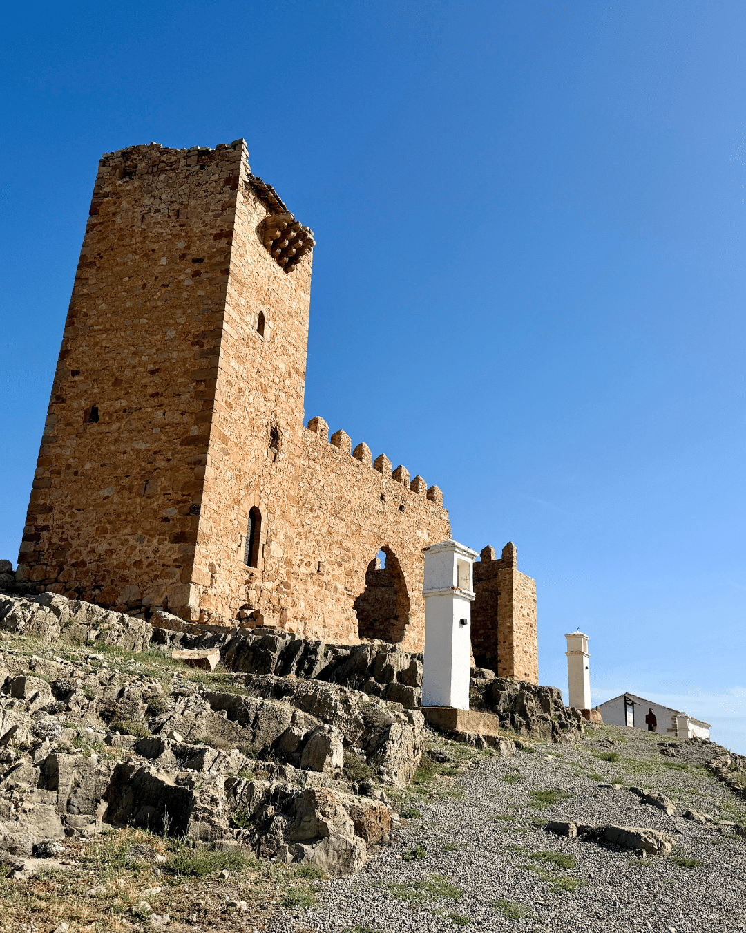 Una antigua fortaleza de piedra con una alta torre cuadrada y muros almenados se asienta en lo alto de una colina rocosa de Asturias bajo un cielo azul despejado, con un camino de grava y balizas blancas que conducen hasta ella.