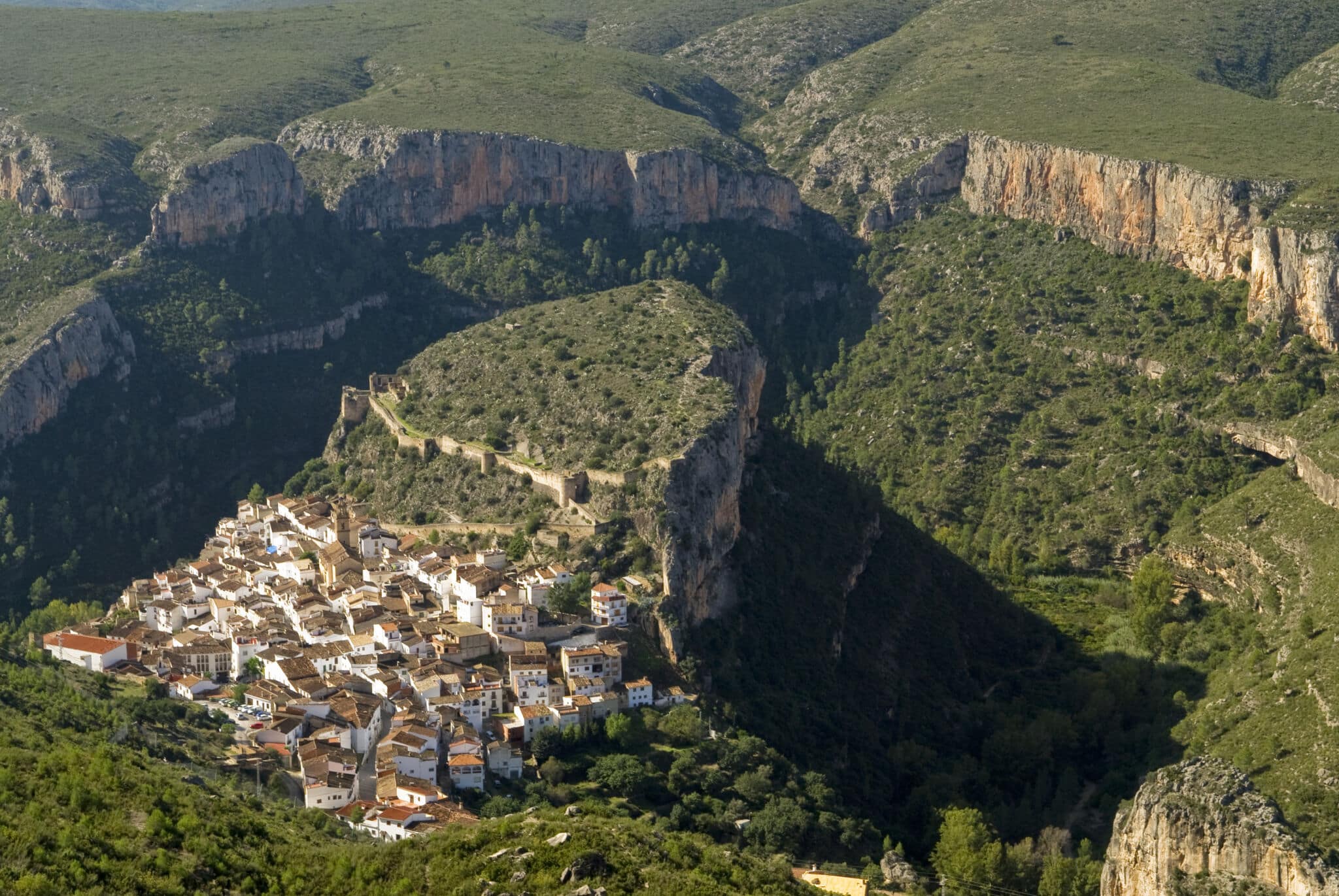 Un pequeño pueblo de casas blancas y tejados de terracota se asienta en la base de escarpadas colinas verdes y dramáticos acantilados rocosos, rodeado de exuberante vegetación y terreno accidentado.