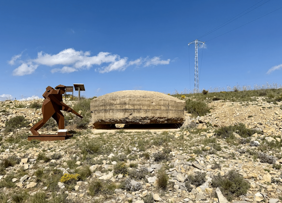 Una escultura metálica de un soldado junto a un búnker de hormigón en un terreno rocoso de Asturias, bajo un cielo azul con nubes. Al fondo se ve una línea eléctrica y una señal informativa.