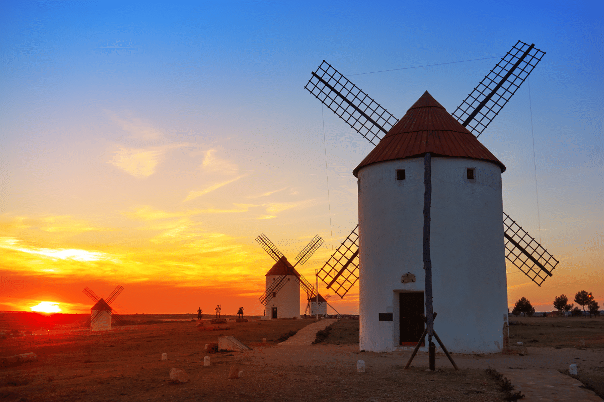 Tres molinos de viento blancos tradicionales con tejados rojos se alzan en una llanura abierta al atardecer, con un vibrante cielo naranja y azul de fondo. El sol está bajo en el horizonte, proyectando una cálida luz sobre la escena.