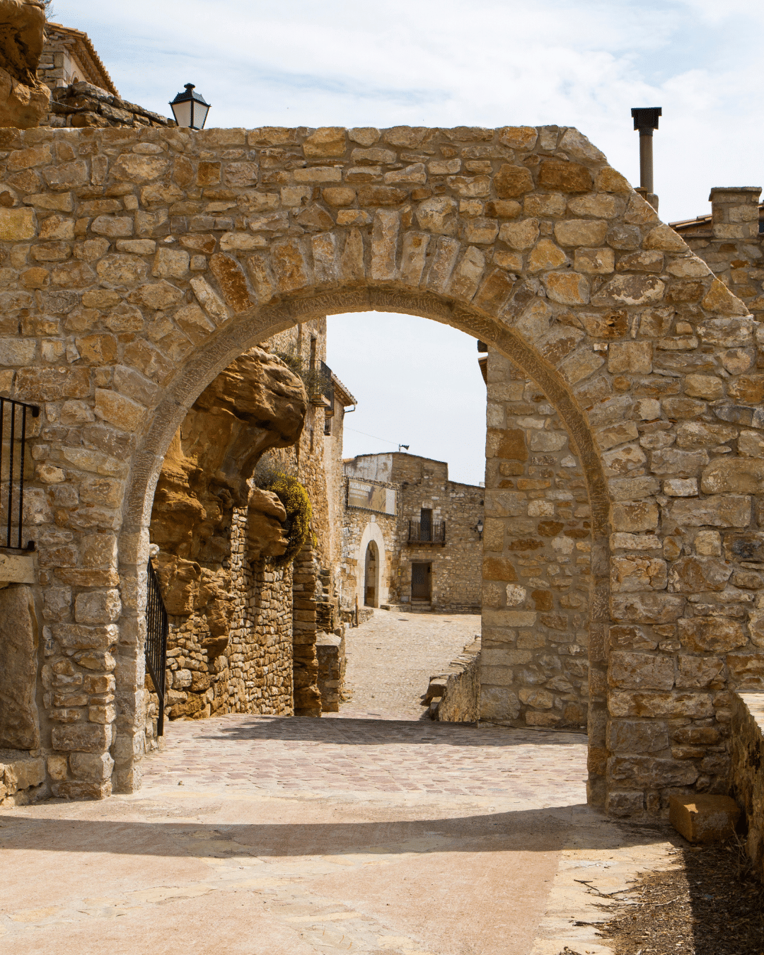 Un arco de piedra conduce a un pintoresco pueblo con rústicos edificios de piedra bajo un cielo parcialmente nublado. El estrecho sendero se curva hacia delante, bordeado por viejos muros y pequeñas ventanas.
