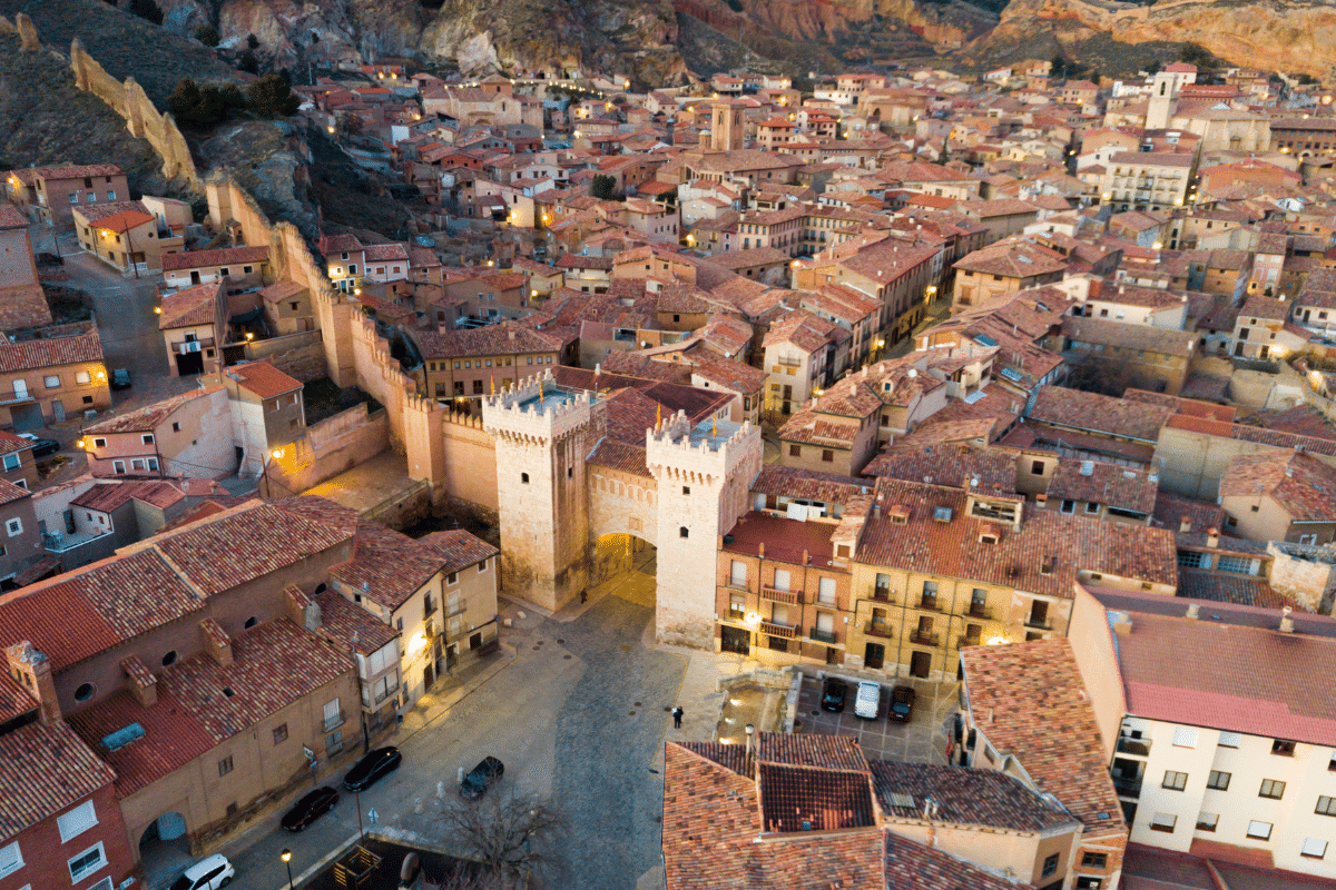 Vista aérea de una histórica ciudad española con tejados de tejas rojas, calles estrechas y una gran puerta de piedra con torres, rodeada de montañas y murallas medievales al atardecer.
