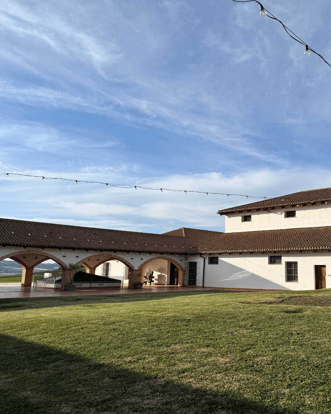 Un gran edificio de estilo español con paredes blancas, tejado de tejas rojas y pasarelas arqueadas evoca el encanto de Asturias bajo un cielo azul, con luces de cuerda en lo alto y un amplio césped que se extiende en primer plano.