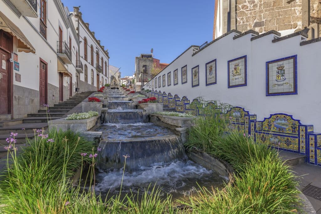 Una cascada de agua fluye por escalones de piedra entre edificios blancos decorados con crestas y azulejos de cerámica de colores, enmarcados por plantas verdes y flores rojas, bajo un cielo azul despejado.