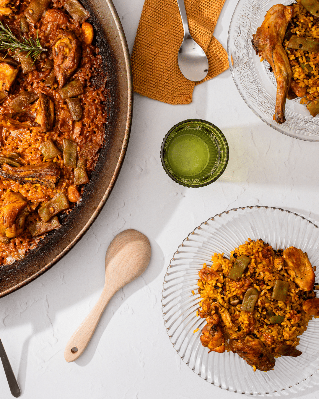 Una sartén con paella de pollo, judías verdes y arroz se encuentra junto a dos platos de cristal con raciones del plato. Una bebida verde, una cuchara de madera y utensilios metálicos están dispuestos sobre una superficie blanca.
