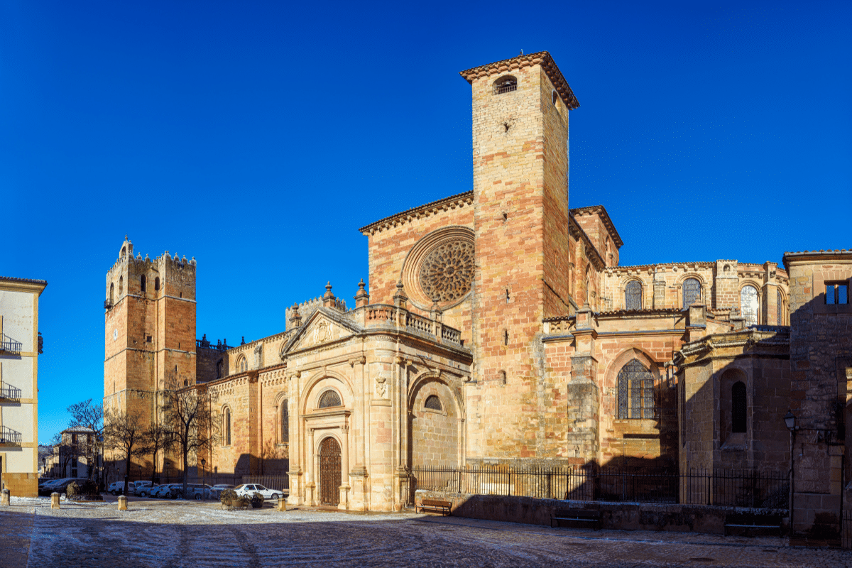 Una gran catedral románico-gótica con rosetones y un alto campanario cuadrado se alza en una plaza iluminada por el sol bajo un cielo azul despejado, con los coches cercanos y algunos edificios a la vista.