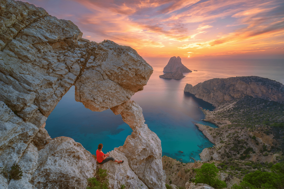 Una persona con camisa roja se sienta en un acantilado rocoso con un arco natural, con vistas al agua turquesa y a una isla lejana al atardecer, con espectaculares nubes naranjas y rosas en el cielo.