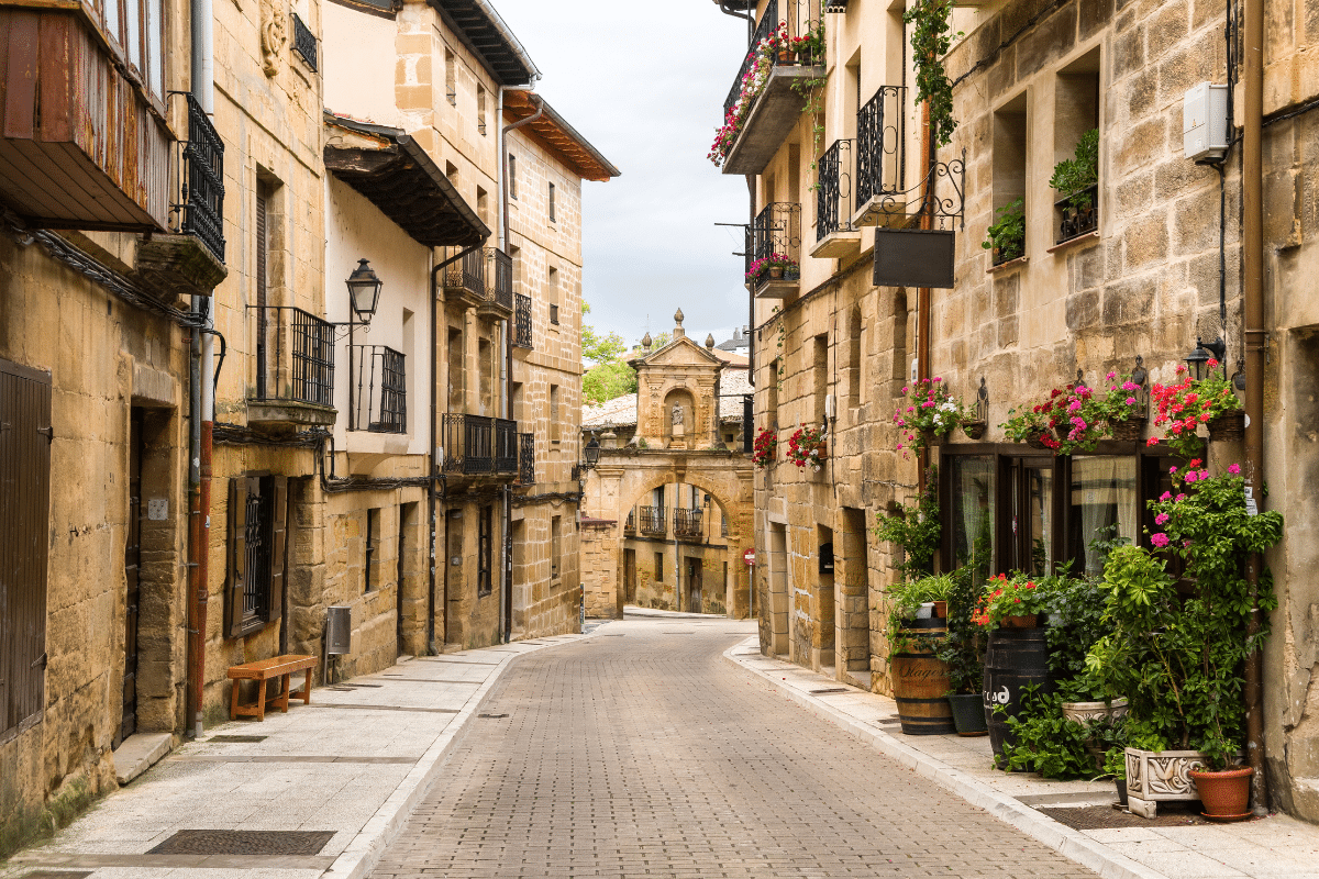 Una calle estrecha y sinuosa de una histórica ciudad europea, flanqueada por viejos edificios de piedra, balcones con macetas de flores y puertas de madera. La calle se curva hacia un arco de piedra al fondo.