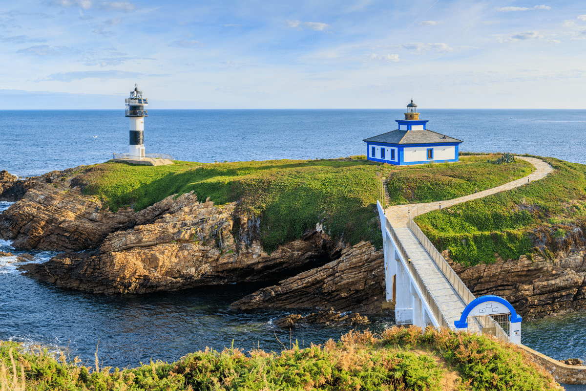 Una vista panorámica de la costa con dos faros sobre acantilados rocosos, conectados por una pasarela blanca sobre el mar. Un faro es alto y rayado, y el otro es pequeño con paredes azules y blancas. El cielo está despejado y azul.