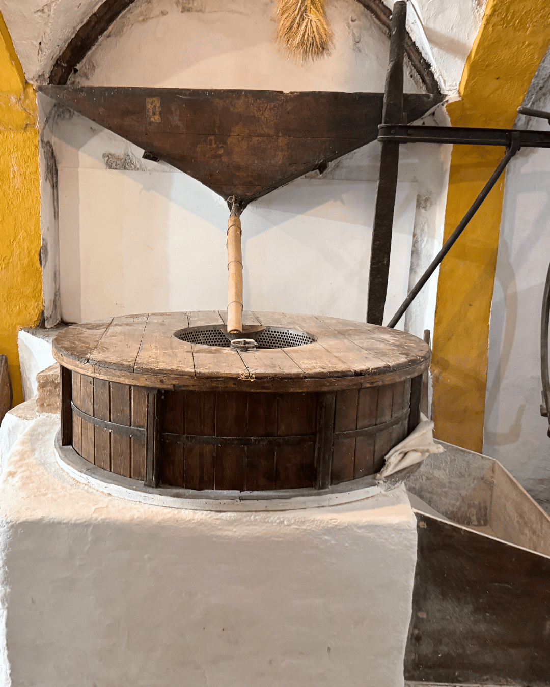 Un molino de madera tradicional de Asturias, con una piedra de moler redonda y cubierta, se asienta sobre una base de piedra blanca, rodeado de maquinaria rústica de madera y metal en una sala con paredes pintadas de amarillo y blanco.