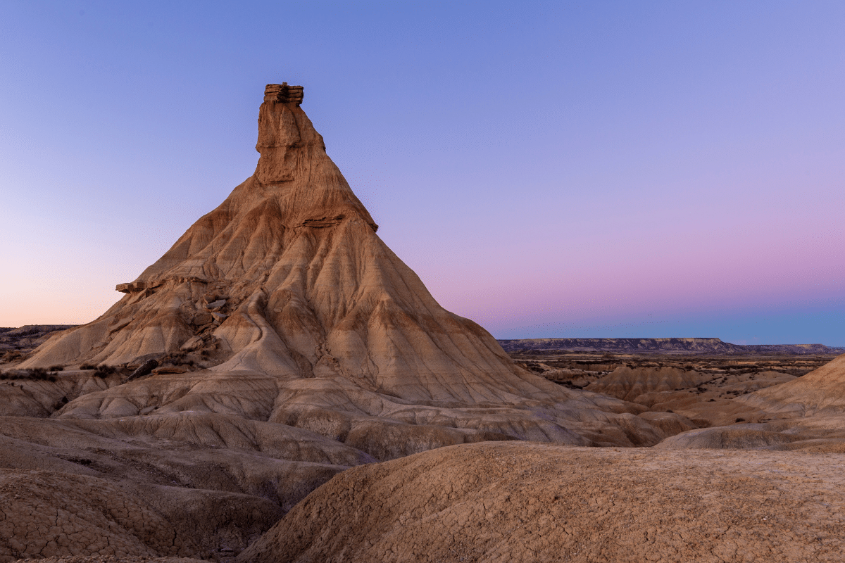 Una escarpada formación de arenisca de forma única se eleva sobre un paisaje árido y rocoso bajo un cielo despejado al atardecer, con suaves tonos rosas y azules en el horizonte.