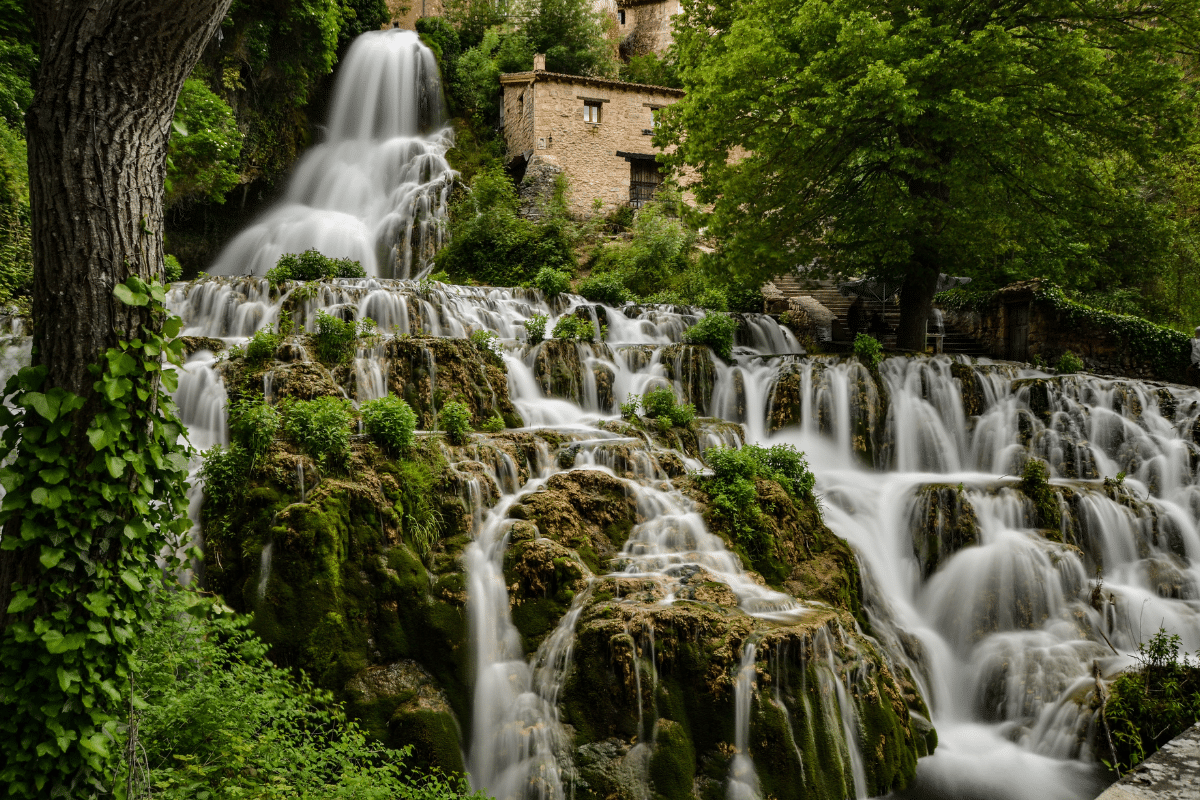 Un exuberante paisaje verde presenta cascadas sobre rocas musgosas, con un viejo edificio de piedra y árboles al fondo. El efecto de larga exposición hace que el agua parezca suave y sedosa.