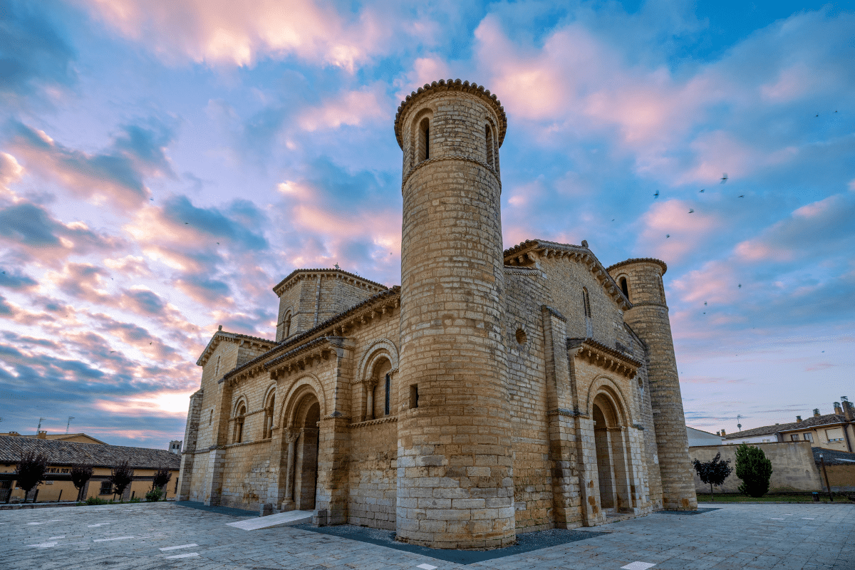 Una iglesia románica de piedra con arcos de medio punto y torres se alza bajo un cielo colorido al atardecer, con nubes rosas y azules. El edificio está rodeado por una zona pavimentada y algunas casas al fondo.