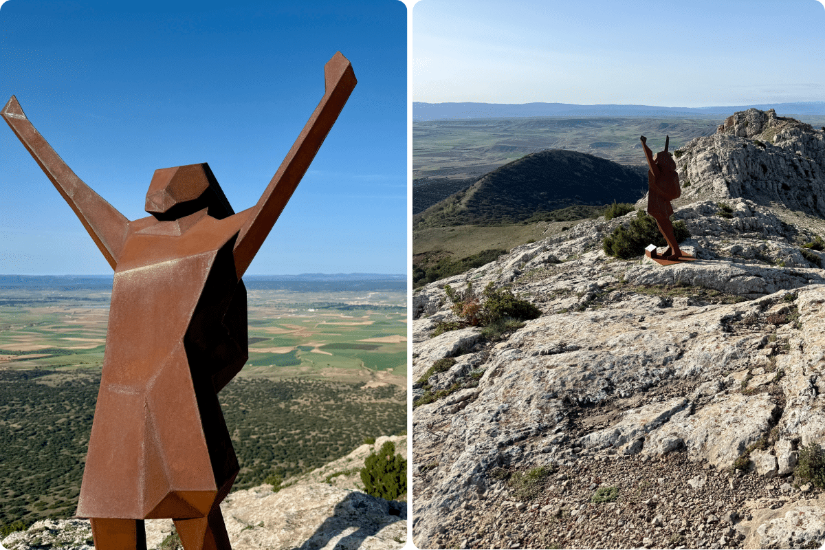 Una escultura geométrica de metal de color óxido de una persona con los brazos levantados se alza en la cima de una colina rocosa en Asturias, con vistas a un vasto paisaje verde bajo un cielo azul despejado.
