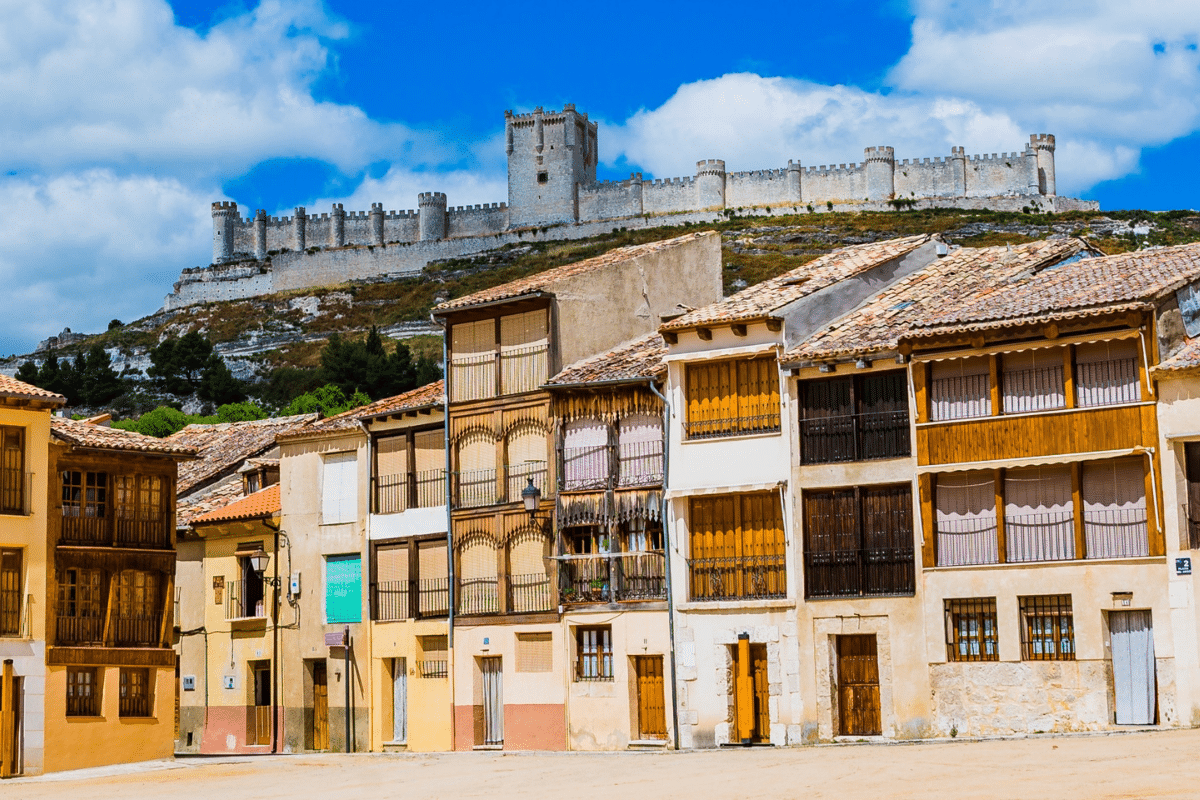 Coloridas casas tradicionales bordean una plaza, con un gran castillo medieval de piedra encaramado en lo alto de una colina al fondo, bajo un cielo azul brillante con nubes dispersas.