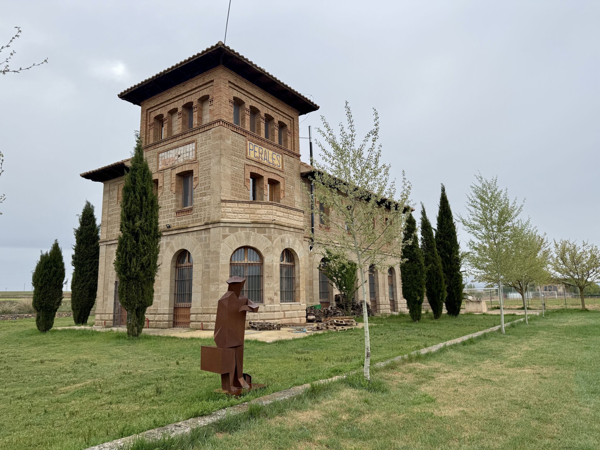 Un edificio de ladrillo y piedra de dos plantas, con ventanas arqueadas y tejado de tejas, se alza sobre un prado herboso en Asturias, con altos cipreses a su lado y una escultura metálica de una persona cargando una maleta en primer plano.