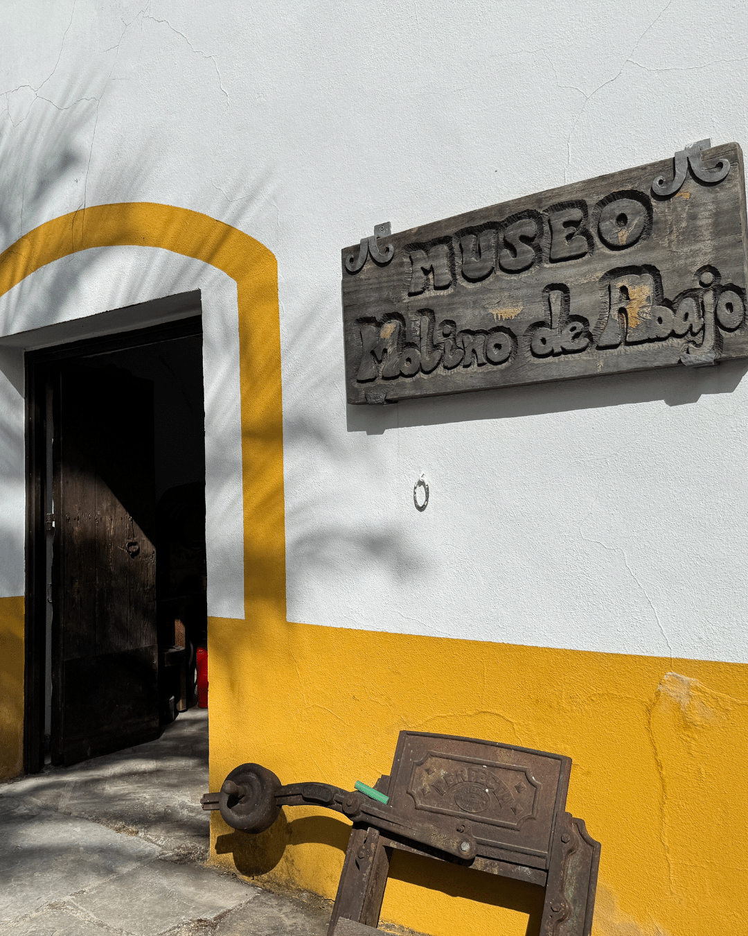 Un rústico cartel de madera en el que se lee Museo Molino de Bajo cuelga de una pared blanca y amarilla sobre una oscura puerta abierta en Asturias; un viejo artefacto metálico descansa cerca sobre el pavimento de piedra iluminado por el sol.