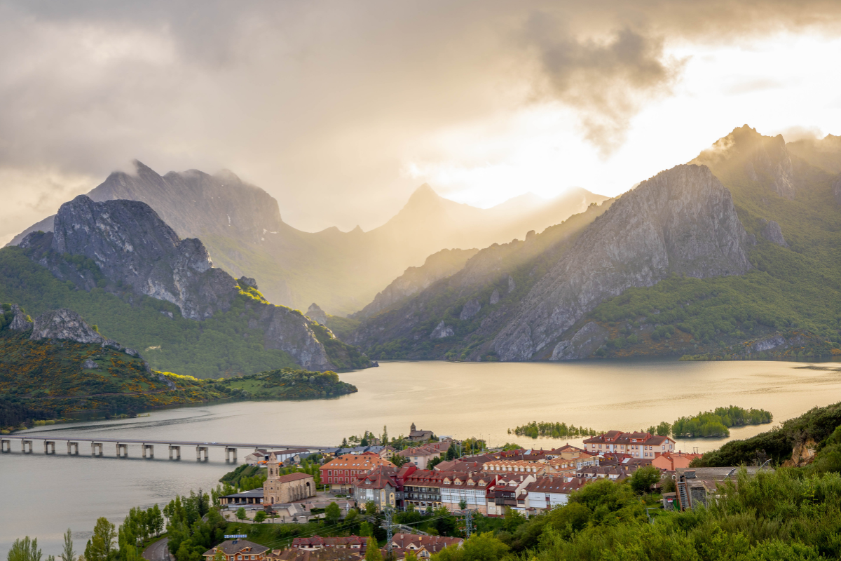 Un pintoresco pueblo a orillas de un lago enclavado entre verdes montañas al atardecer, con la luz del sol atravesando las nubes y un largo puente que cruza el agua. Edificios de tejados rojos se agrupan cerca de la orilla.