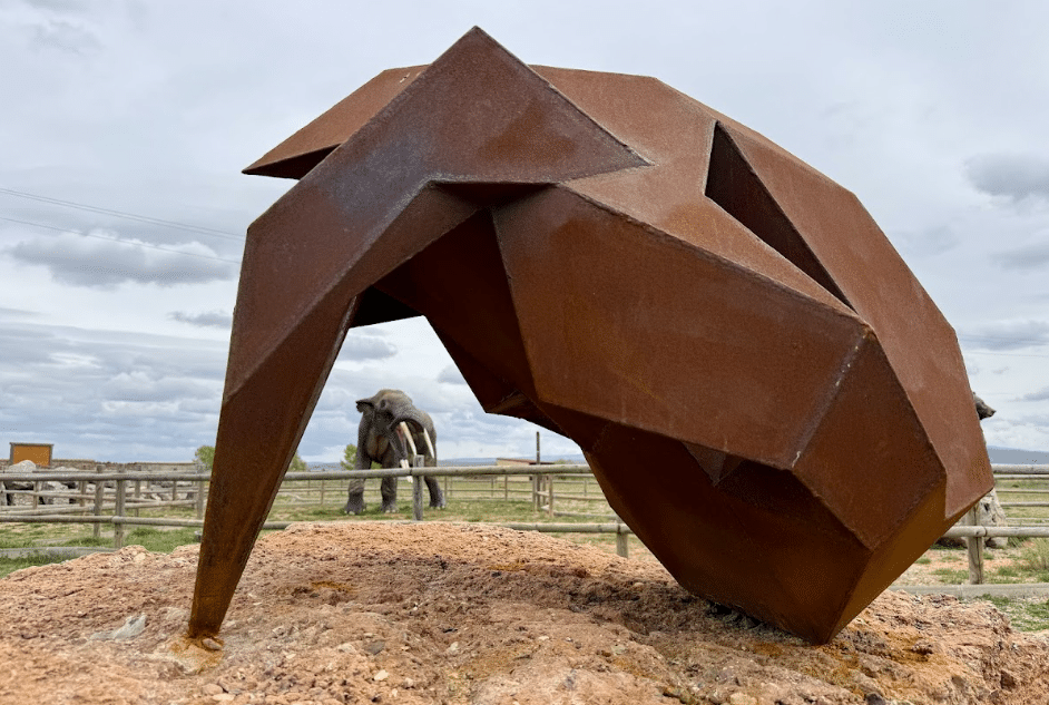 Una escultura de metal oxidado y anguloso se asienta sobre una roca en primer plano, con una estatua de elefante de tamaño natural visible al fondo, situada en una zona vallada al aire libre bajo el cielo nublado de Asturias.