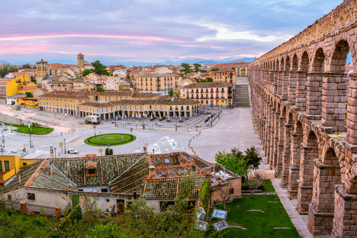Vista de Segovia, España, con el antiguo acueducto romano a la derecha, edificios tradicionales con tejados de arcilla, una rotonda con vegetación y el paisaje urbano bajo un colorido cielo al atardecer.