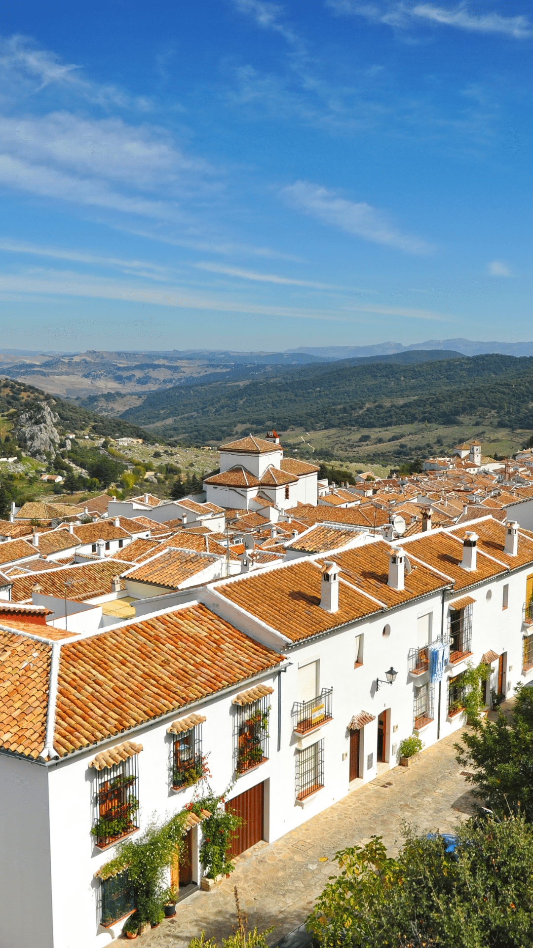 Casas blancas con tejados de terracota se alinean en la ladera de un pueblo de Asturias, bajo un cielo azul brillante y rodeado de verdes colinas y montañas lejanas.