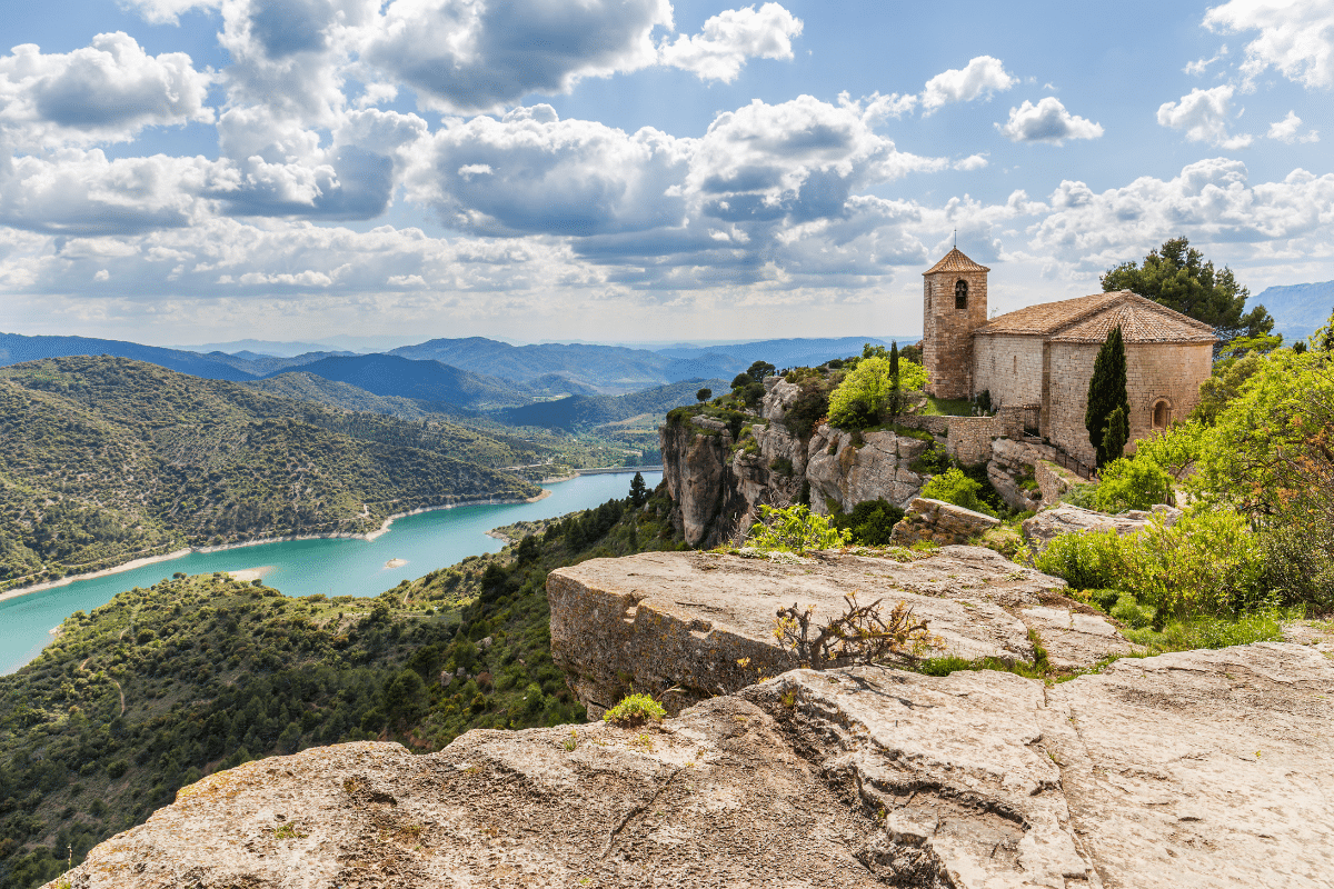 Una pequeña iglesia de piedra se asienta sobre un acantilado rocoso con vistas a un serpenteante río turquesa y a verdes colinas bajo un cielo parcialmente nublado. El paisaje es exuberante y amplio, con montañas lejanas en el horizonte.