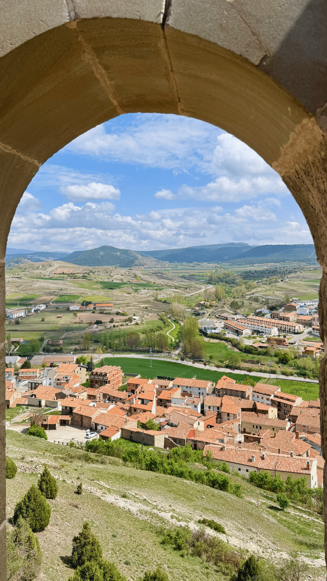 Vista panorámica de un pueblo con tejados de tejas rojas, campos verdes y colinas lejanas, vista a través de un arco de piedra bajo un cielo parcialmente nublado.