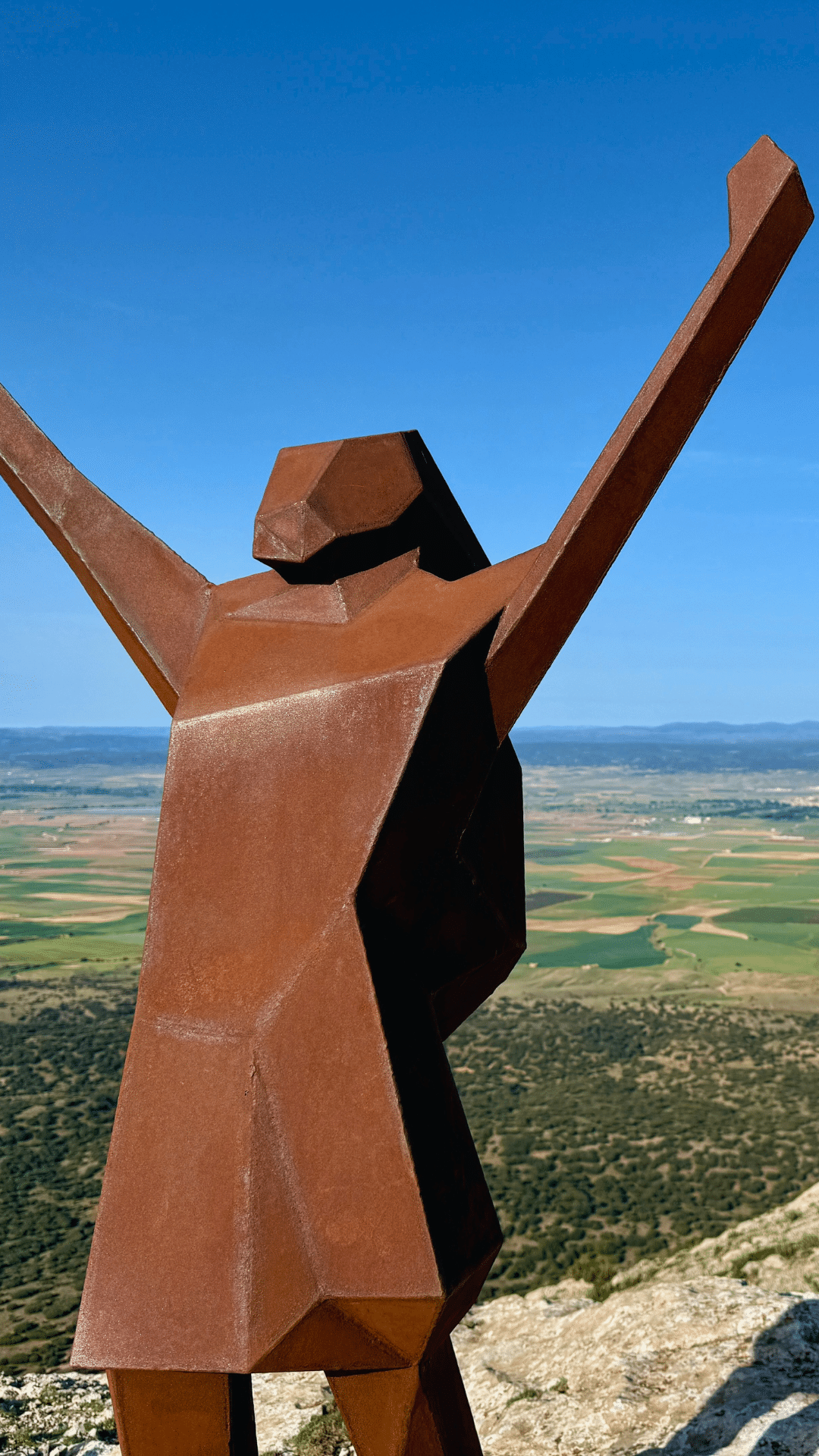 Una escultura metálica oxidada y angulosa de una figura humana con los brazos levantados se alza en lo alto de una colina, dominando un vasto paisaje de campos verdes y montañas lejanas bajo un cielo azul despejado.