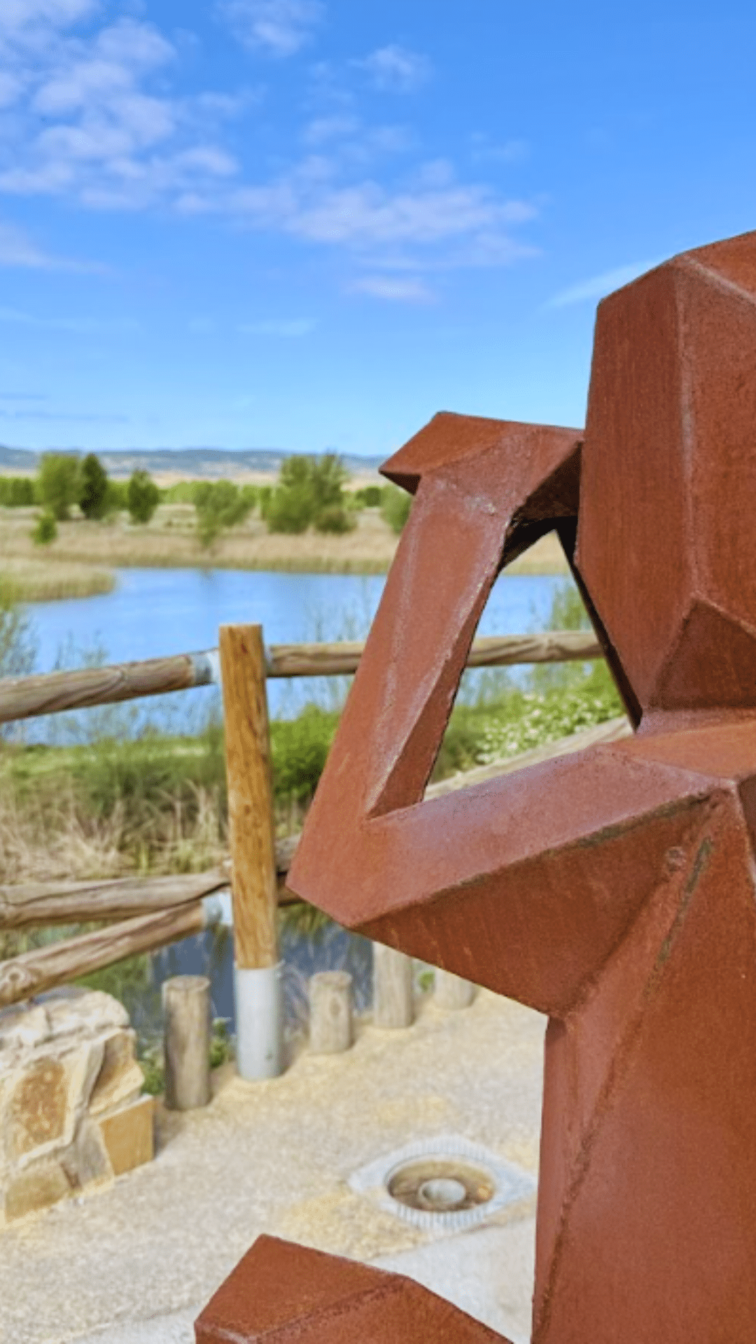 Primer plano de una escultura de metal oxidado y anguloso que recuerda a un mono, colocada al aire libre junto a una valla de madera, con un estanque, vegetación y un cielo azul de fondo.