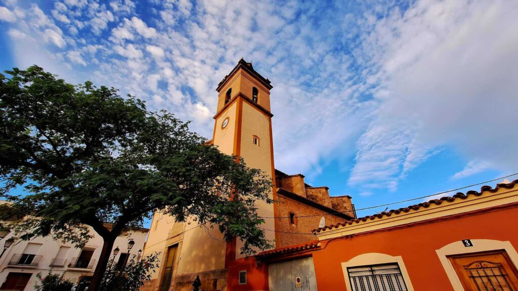 Una alta torre de iglesia con un reloj se alza sobre un cielo azul vibrante con nubes blancas dispersas, rodeada de árboles y edificios vecinos de colores.