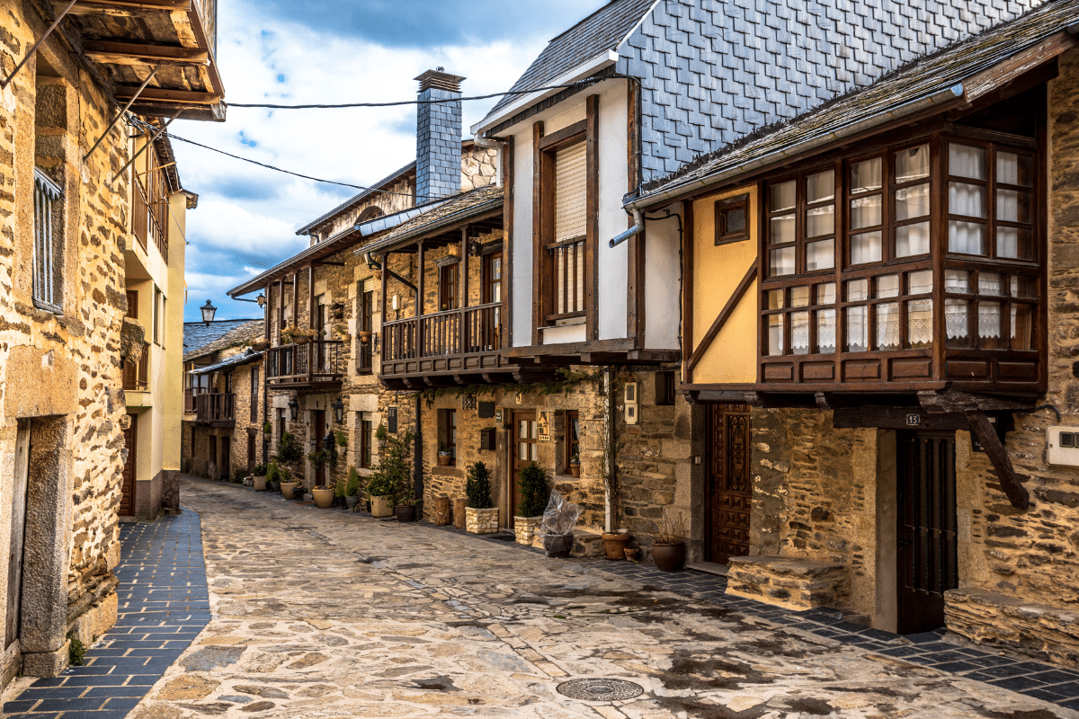 Una estrecha calle adoquinada bordeada de rústicas casas de piedra con balcones de madera, macetas y ventanas enrejadas bajo un cielo nublado en un pintoresco pueblo europeo.