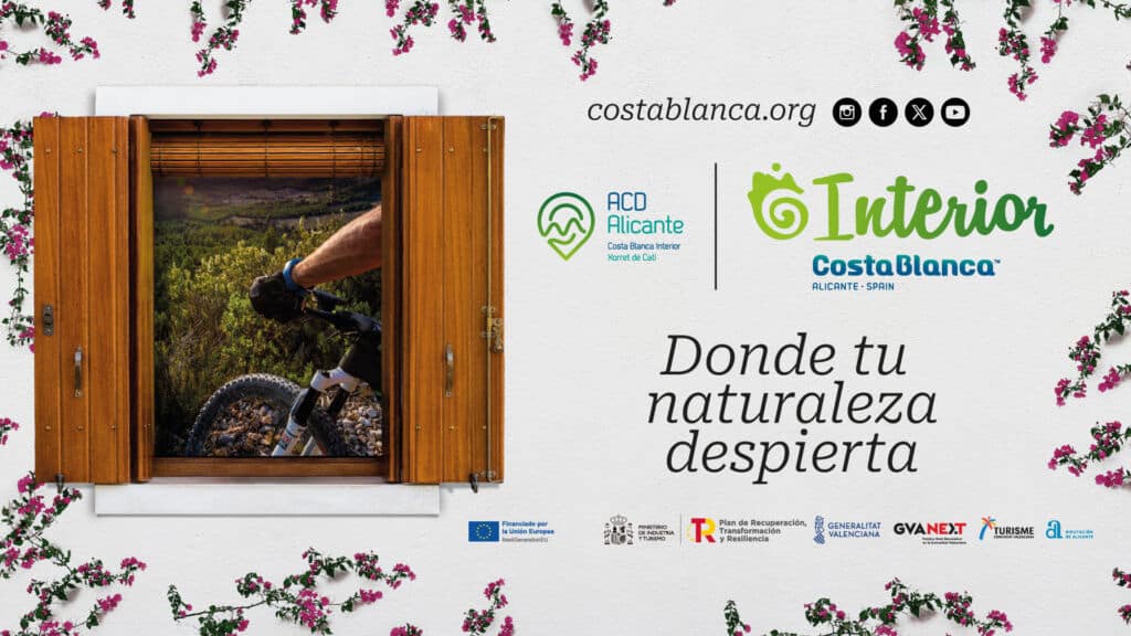 Una ventana de madera enmarca a un ciclista que atraviesa un paisaje natural. Flores rosas bordean la ventana. A la derecha, logotipos y texto: "Interior Costa Blanca. Donde tu naturaleza despierta".