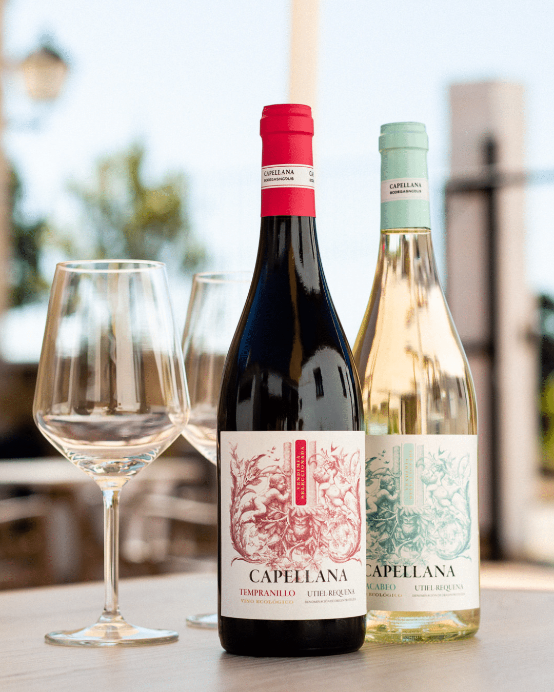 Dos botellas de vino Capellana, una roja y otra blanca, están sobre una mesa junto a dos copas vacías. Las etiquetas presentan diseños intrincados. El fondo está suavemente difuminado con elementos exteriores visibles.