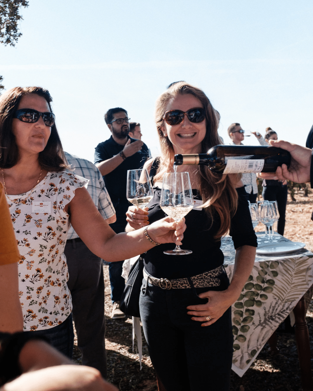 Dos mujeres con gafas de sol sonríen mientras una de ellas se sirve vino en su copa en una reunión al aire libre. Al fondo, otras personas charlan bajo un cielo despejado.