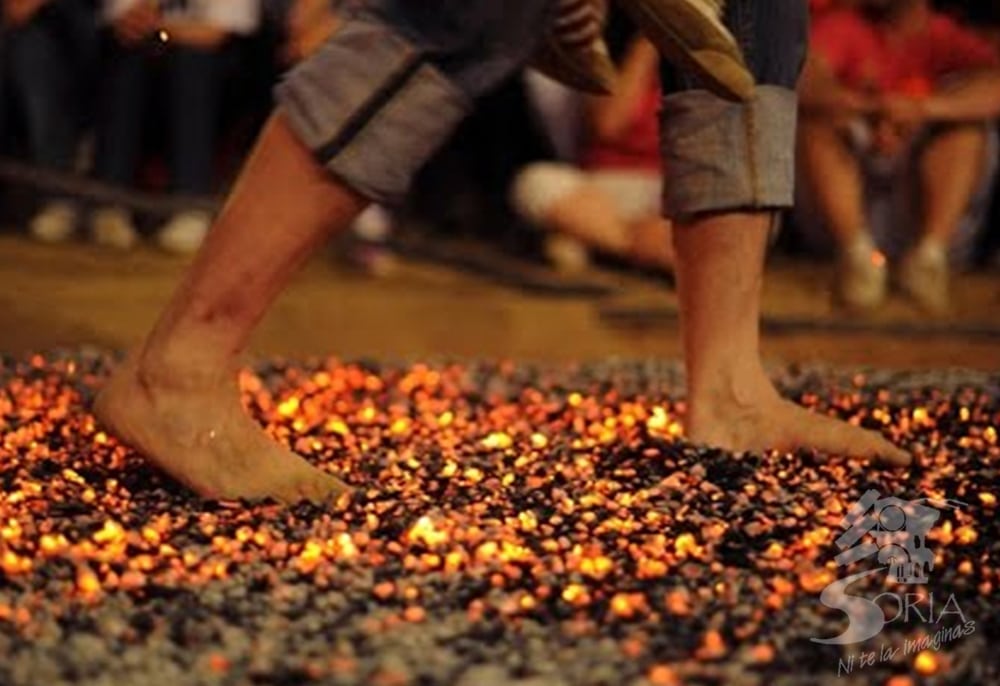 Pies descalzos caminando sobre brasas ardientes durante un evento de "firewalking", con espectadores difuminados en el fondo. La escena es intensa y pone de relieve la atrevida actividad.