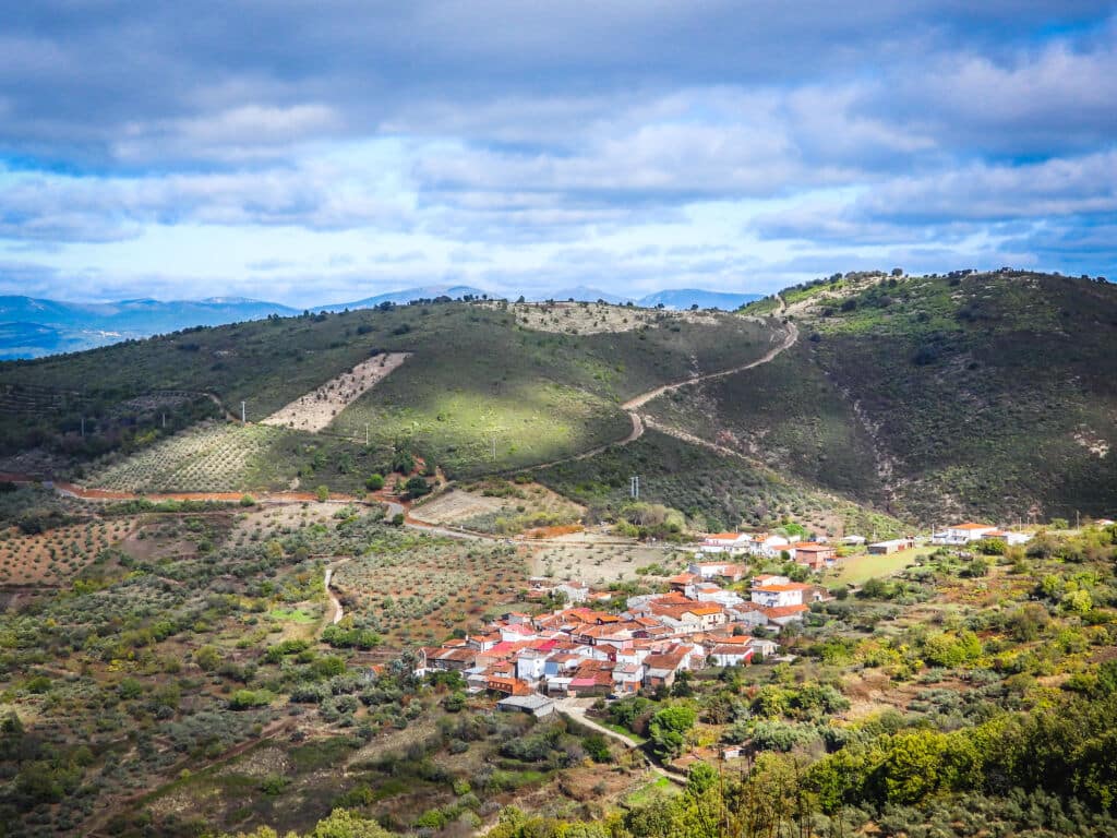 Un pequeño pueblo con casas de tejados rojos se alza entre verdes colinas y tierras de cultivo bajo un cielo parcialmente nublado, con zonas iluminadas por el sol que resaltan algunos rincones del paisaje.