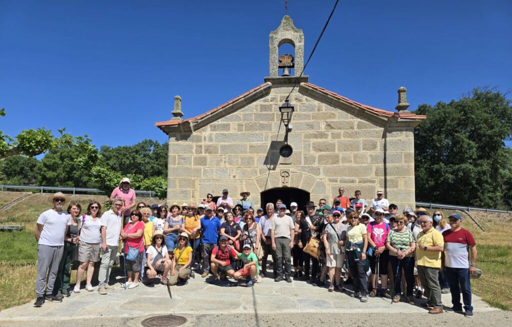 Un numeroso grupo de personas posa para una foto frente a una pequeña capilla de piedra con un campanario en un día soleado. El grupo está formado por adultos y niños, la mayoría de los cuales llevan gorras y ropa informal. Al fondo se ven árboles y el cielo azul.