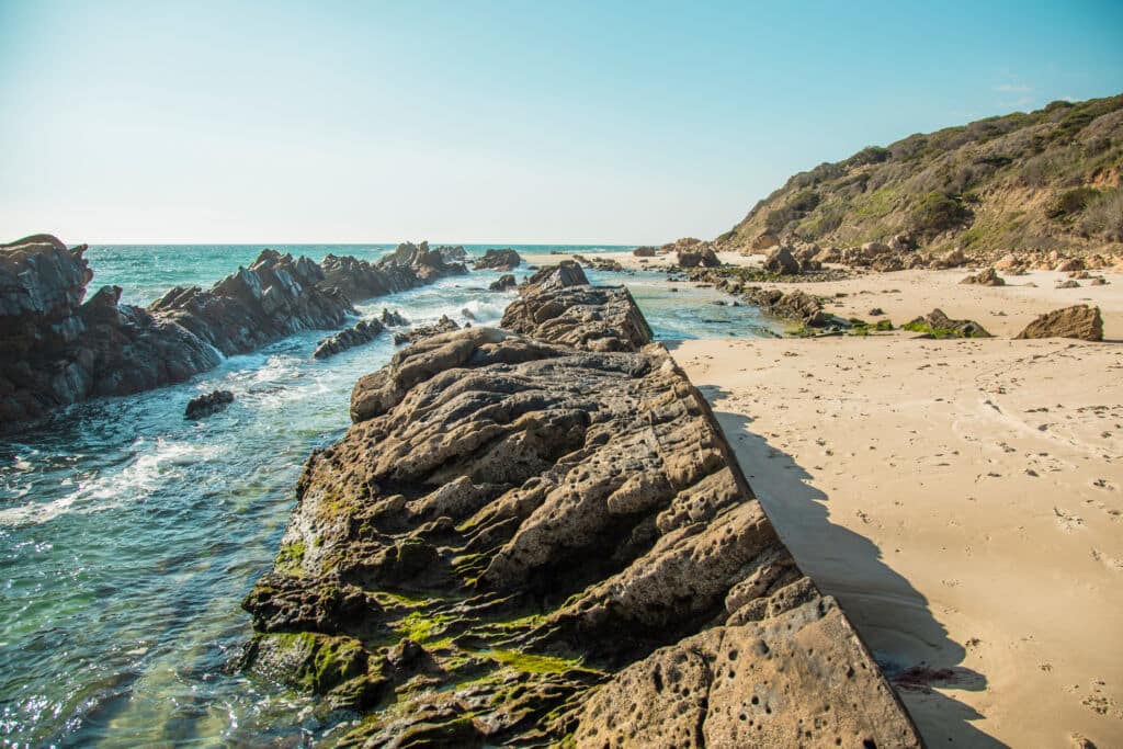 Los afloramientos rocosos se adentran desde la playa de arena hacia el océano de color turquesa, donde las suaves olas rompen contra las rocas. A la derecha, bajo un cielo azul claro, se alza una ladera verde cubierta de matorrales.