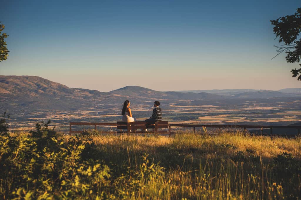 Dos personas están sentadas en un banco con vistas a un valle pintoresco, con montañas a lo lejos bajo un cielo azul y despejado, rodeadas de hierba y árboles bañados por el sol.