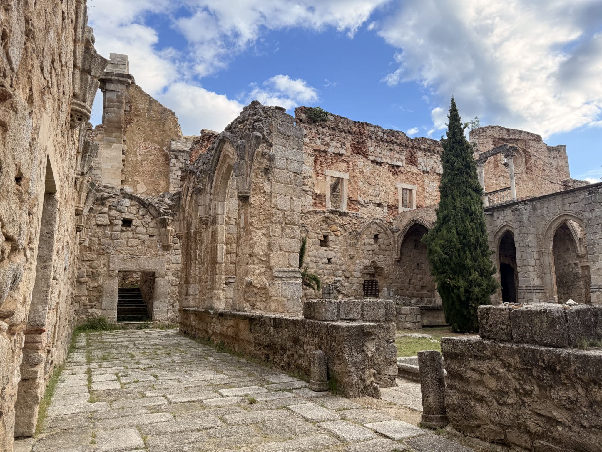 Antiguas ruinas de piedra con arcos y muros erosionados en Asturias, un alto ciprés y parches de hierba bajo un cielo azul parcialmente nublado.