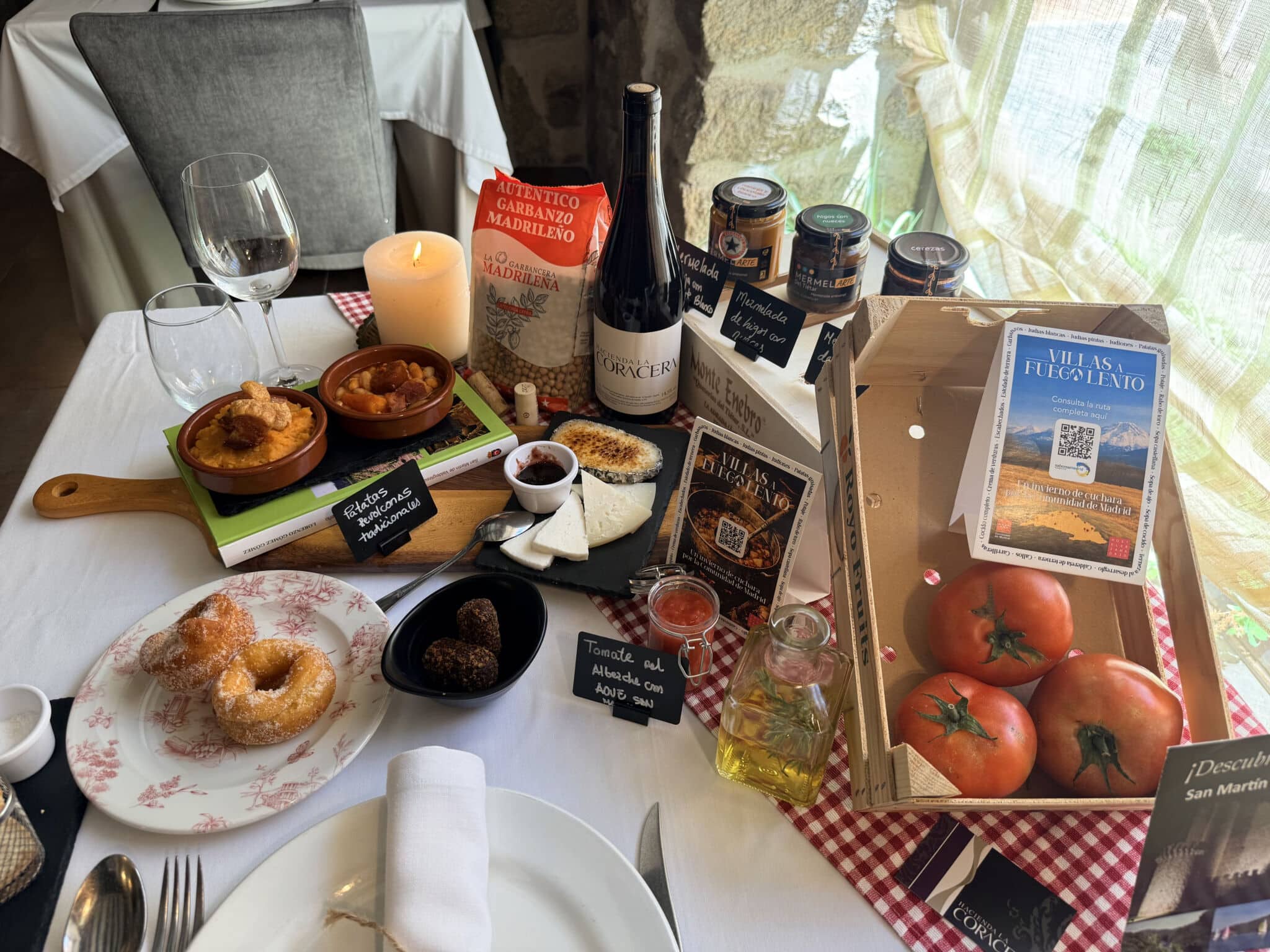 Una mesa con vino, queso, pan, bollería, tomates, aceite de oliva y diversos alimentos españoles de Asturias, junto a libros de cocina, una vela y una decoración rústica, crean un ambiente acogedor y agradable.