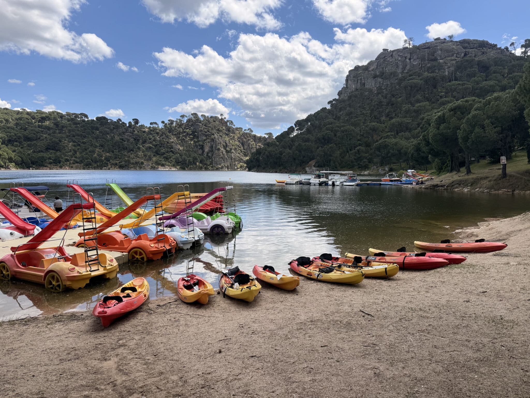 Coloridos kayaks y barcas de pedales se alinean en la orilla de un lago de arena en Asturias, con aguas tranquilas, colinas, árboles y cielos parcialmente nublados de fondo. Al otro lado del agua, cerca de una colina rocosa, se ven otras embarcaciones.