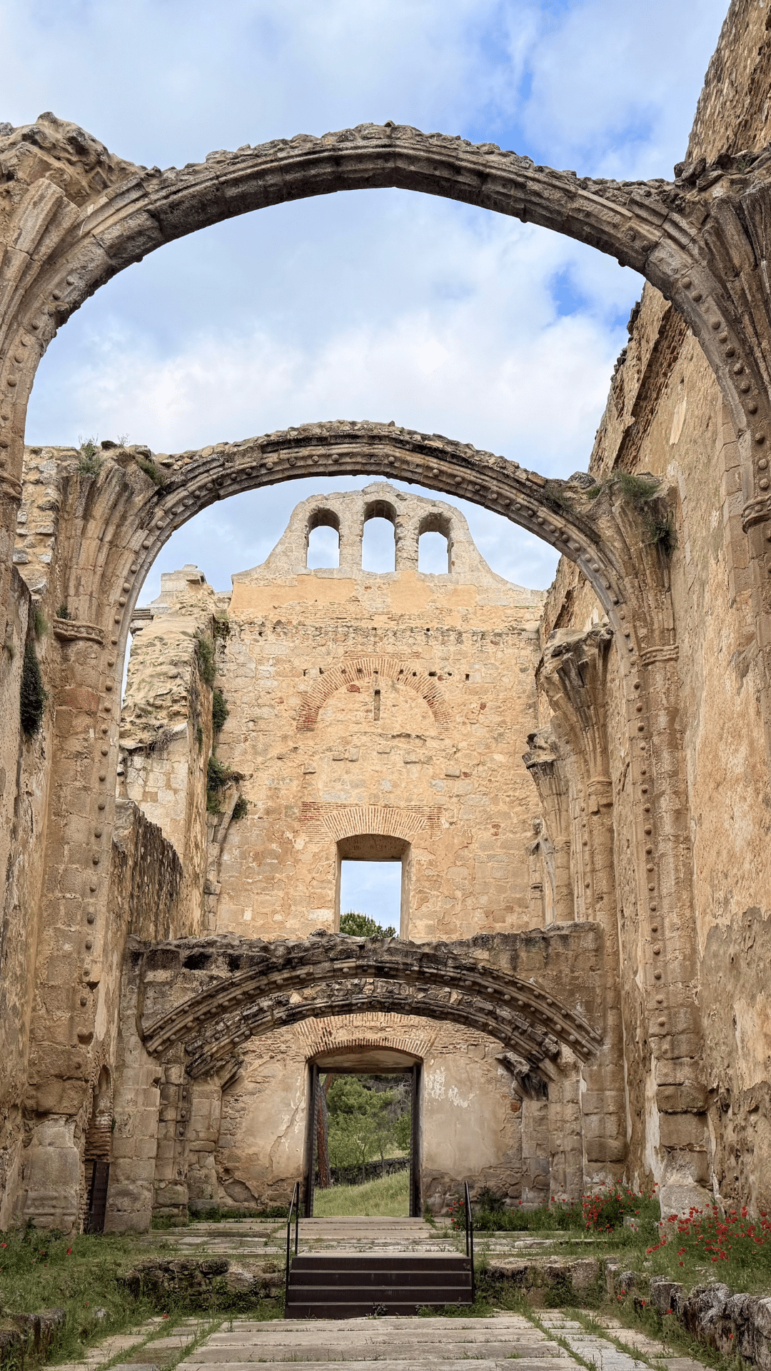 Ruinas de un antiguo edificio de piedra con grandes arcos, un tejado parcialmente derruido y tres huecos para campanas en la parte superior, enclavado en la exuberante vegetación de Asturias bajo un cielo parcialmente nublado.