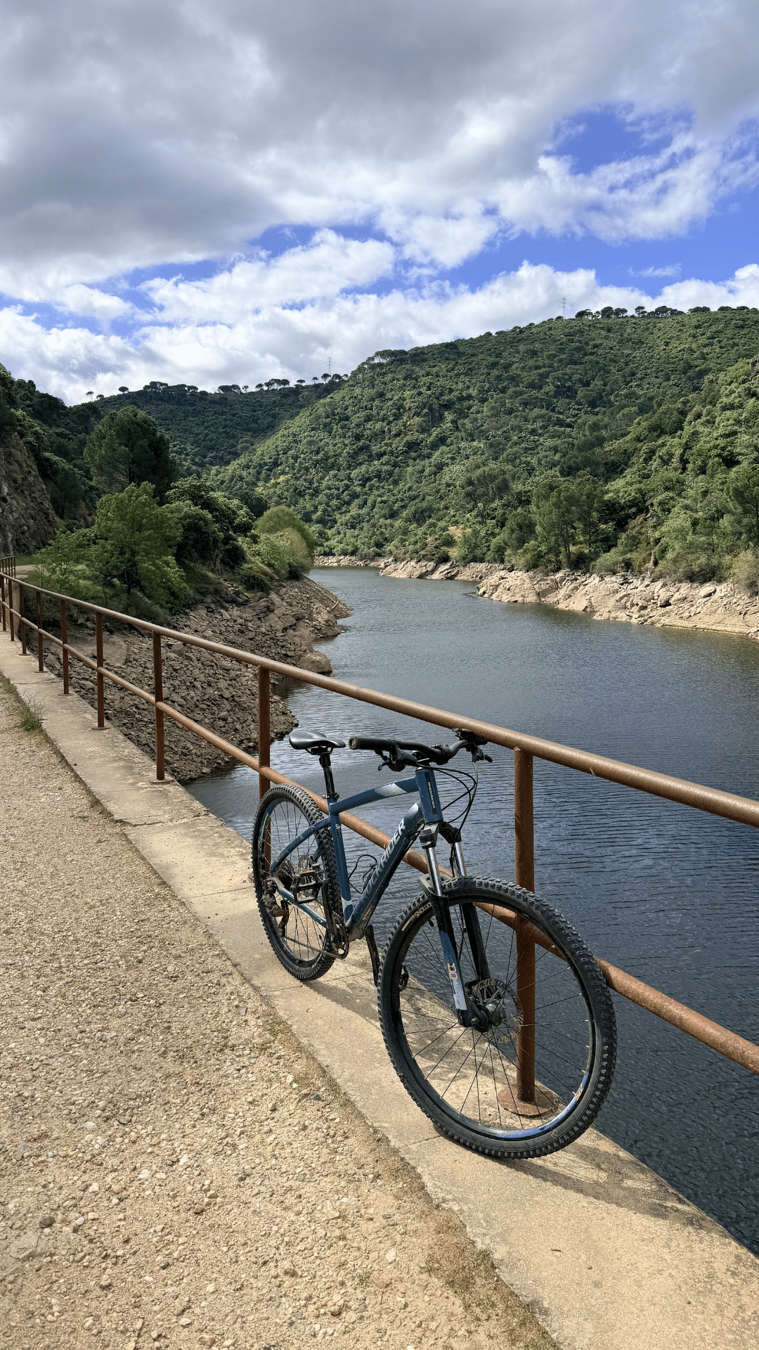 Una bicicleta de montaña descansa contra una barandilla metálica en un sendero junto a un río en Asturias, rodeada de verdes colinas y árboles bajo un cielo parcialmente nublado.