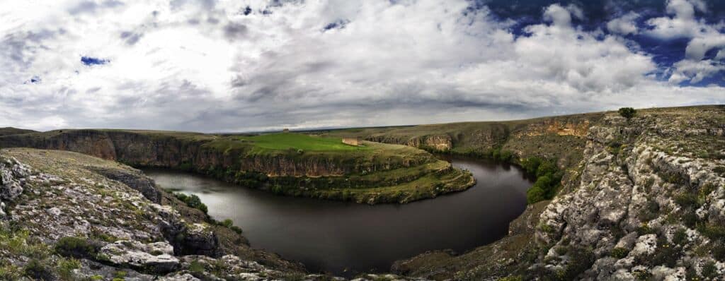 Un río ancho serpentea alrededor de una meseta verde en lo alto de una colina, rodeada de acantilados rocosos bajo un cielo nublado y espectacular, creando un paisaje pintoresco y escarpado.