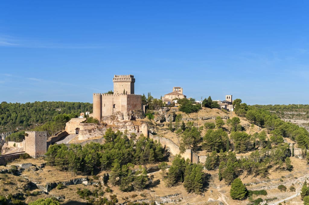 Un castillo medieval con una alta torre central se alza sobre una colina rodeada de árboles y edificios históricos, bajo un cielo azul y despejado.