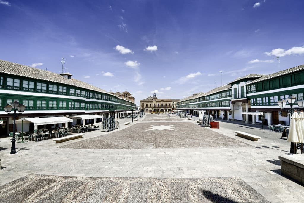 Una plaza amplia y diáfana con balcones de madera de color verde a ambos lados, suelo de adoquines, mesas al aire libre y un edificio al fondo, bajo un cielo azul brillante salpicado de nubes.
