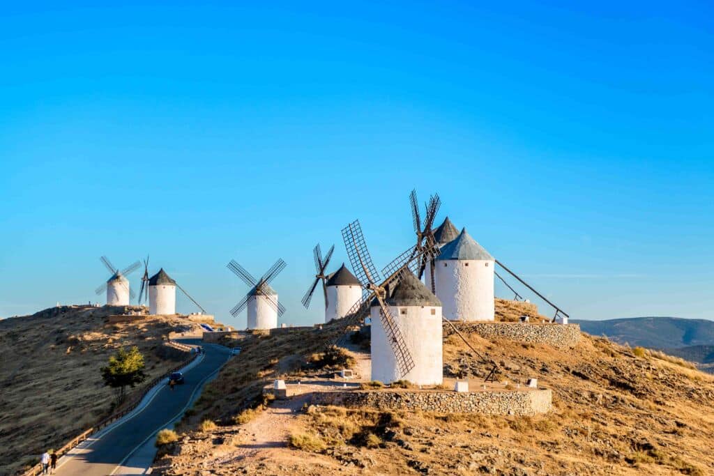 Seis molinos de viento blancos tradicionales con tejados oscuros se alzan en un paisaje seco y montañoso bajo un cielo azul y despejado. Una carretera sinuosa discurre junto a los molinos.