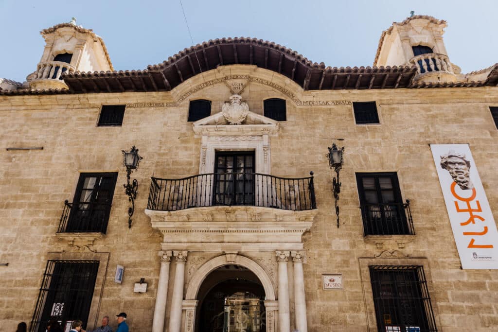 Edificio histórico de piedra con entrada en arco, dos torres, balcón y un cartel del museo en el que se lee «MUSEO», junto a la imagen de una estatua. Se aprecian detalles ornamentados sobre la puerta principal. El cielo está despejado.