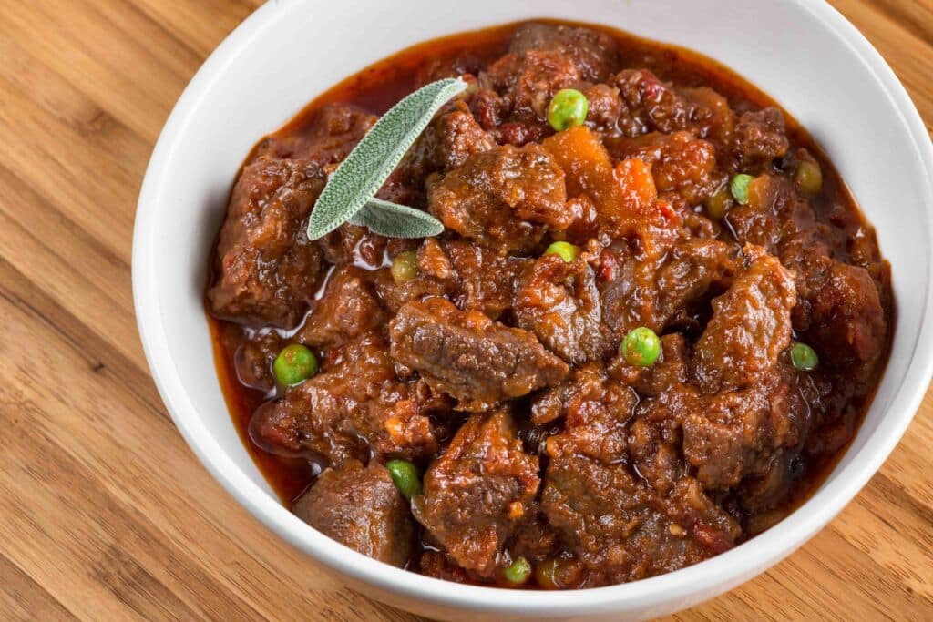 Sobre una superficie de madera hay un cuenco blanco lleno de estofado de ternera con trozos de carne, guisantes y zanahorias en una salsa roja espesa, adornado con hojas frescas de salvia.